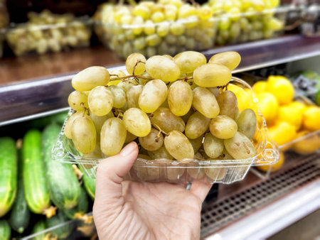 A person holds a clear container filled with fresh green grapes in a grocery store, surrounded by colorful fruits and vegetables.の写真素材