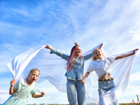 Three children joyfully playing outdoors with flowing fabric under a bright blue sky, enjoying a breezy day in an open fieldの写真素材