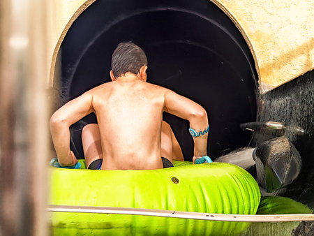 A young boy sits on a vibrant green inflatable tube, ready to slide down into a dark water tunnel at an amusement park attraction.の写真素材