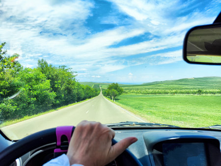 Traveling along a winding road, a hand on the steering wheel, with vibrant green hills and a bright blue sky ahead.の写真素材
