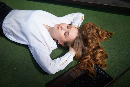 Relaxing outdoors, a young woman lounges on green grass with a laptop nearby on a sunny dayの写真素材