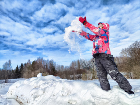 A child dressed in winter clothing joyfully throws snow into the air while standing on a snowy mound in a winter wonderland.の写真素材