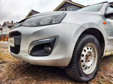 A silver car is parked on damp ground surrounded by houses on a cloudy day, showing its front and...