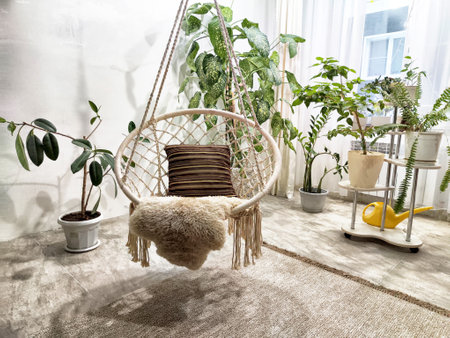 A macrame hanging chair with a plush cushion sits amid thriving indoor plants in a bright, sunny room filled with natural light.の写真素材