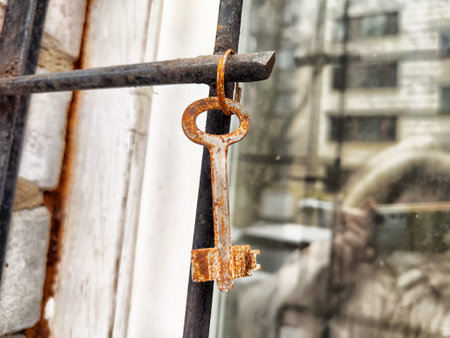 A weathered key with rust lies on a metal hook beside a window frame, reflecting the urban environment in soft afternoon light.の写真素材