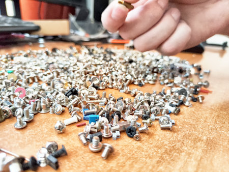 A hand reaches for a distinct screw among a vast assortment of metal fasteners spread across a...