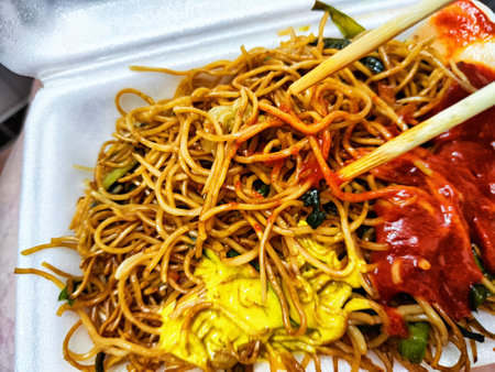 Chow mein noodles topped with red sauce and yellow condiment are being held with chopsticks from a...