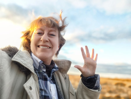 A middle-aged woman with blonde hair smiles brightly while waving her hand outdoors, enjoying a moment of joy in her travel adventure.の写真素材