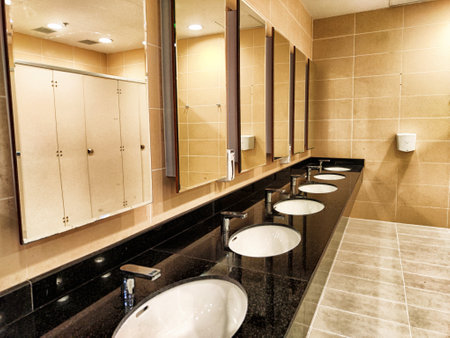 A spacious restroom features several sinks and mirrors, showing a clean design with beige tiles and stainless fixtures.の写真素材
