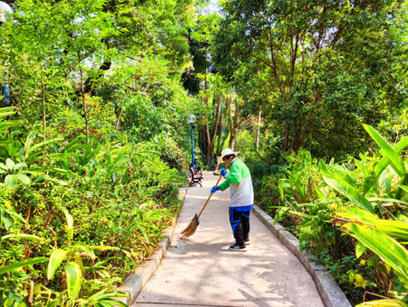 In a vibrant park, a worker diligently sweeps a concrete walkway surrounded by dense greenery and sunny skies.の写真素材