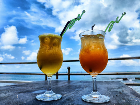 Two vibrant drinks sit on a table overlooking the ocean, with a clear blue sky and fluffy clouds...