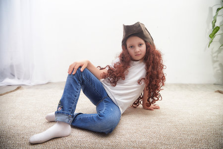 Young child with curly hair and a hat poses playfully on a soft rug in a bright indoor spaceの写真素材