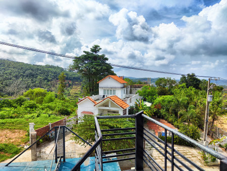 A cozy house with a red roof sits amid vibrant green hills, while dramatic clouds form overhead, creating a picturesque landscape.の写真素材