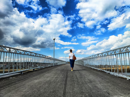 A lone individual strolls along a broad pathway, surrounded by expansive skies filled with clouds and open fields under a bright sun.の写真素材