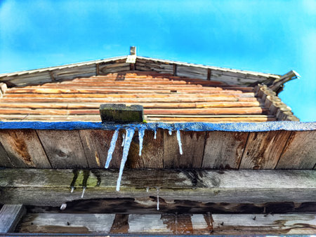 Icicles are forming from the edge of a wooden cabin eave with a vibrant, clear blue sky as the backdrop in winter.の写真素材