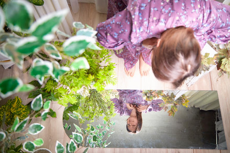 A girl in a floral dress gazes into a mirror surrounded by lush greenery indoors during daylight...