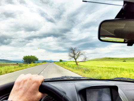 A hand grips the steering wheel as the car moves along a winding road surrounded by vibrant green fields and moody skies.の写真素材