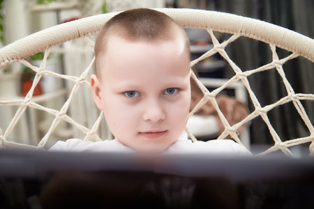 Child focused on a screen while sitting in a macrame chair at home during the afternoonの写真素材