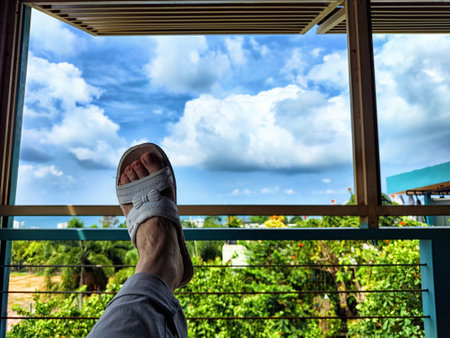 A person relaxes on a balcony, gazing at a vibrant landscape filled with greenery under a bright blue sky with fluffy clouds.の写真素材