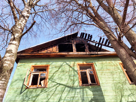 An old green house shows signs of neglect with a collapsing roof and broken windows while trees stand nearby under bright daylight.の写真素材