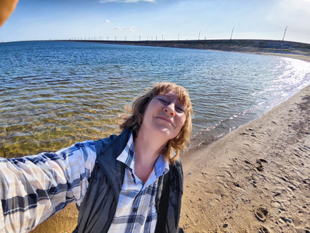 Blonde woman enjoys a sunny day by the water, capturing a joyful selfie while on vacation at the beach, surrounded by nature.の写真素材