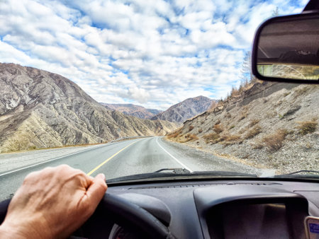 A hand grips the steering wheel while navigating a winding road between rugged mountains, surrounded by a vast sky with clouds.の写真素材
