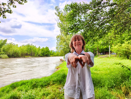 Blonde woman smiles and poses with her hands by a riverbank, enjoying a sunny day surrounded by greenery and tranquility.の写真素材