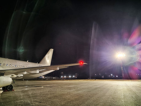 An airplane wing is visible in the forefront at an airport during night time, with floodlights brightly illuminating the tarmac area.の写真素材