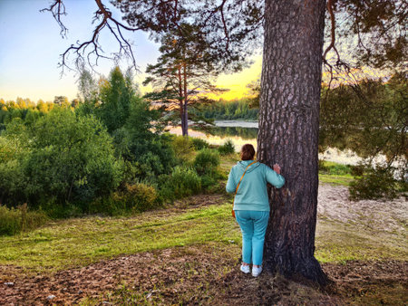A woman in a turquoise outfit stands by a tall tree near a serene lakeside, absorbing the calm beauty of the sunset and nature.の写真素材