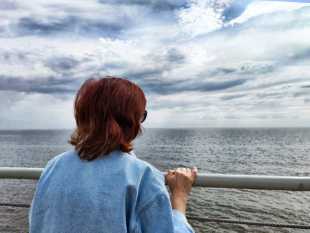 A woman with red hair leans against a railing, looking out at the calm ocean and overcast sky on a tranquil day.の写真素材