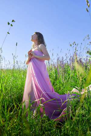 Female model in pink dress standing in green field under blue sky during sunny day capturing natural beauty and serenityの写真素材