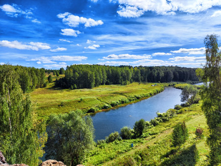 A peaceful river winds through vibrant green fields surrounded by trees, showcasing a clear blue sky with fluffy white clouds.の写真素材