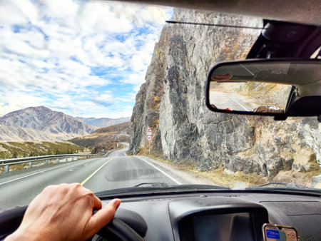 A driver navigates a twisting road bordered by steep rocky cliffs and scenic mountain views under an expansive sky with scattered clouds.の写真素材