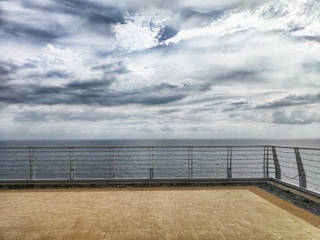 A breathtaking coastal panorama features a gray sky filled with clouds, overlooking a tranquil ocean from a modern terrace.の写真素材