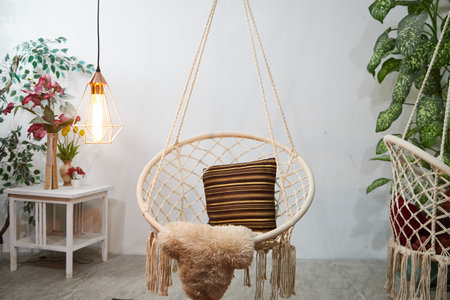 A hammock chair with a striped pillow is suspended in a plush room, surrounded by potted plants and a stylish light fixture, creating a warm, inviting atmosphere perfect for relaxation.の写真素材