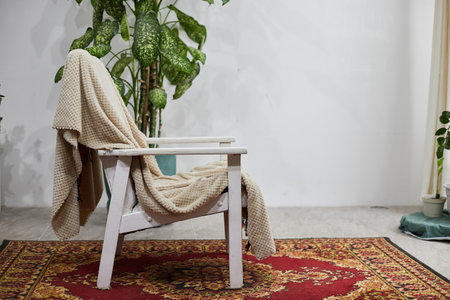 A white wooden chair holds a beige blanket, creating a warm and inviting atmosphere. Surrounding plants add a touch of nature, enhancing the cozy indoor environment.の写真素材