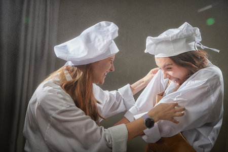Two female friends laugh and playfully toss flour while dressed as chefs, enjoying a lively moment during their fun cooking photoshoot indoorsの写真素材