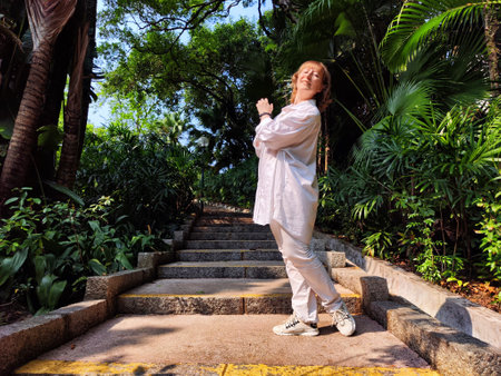 Blonde woman stands on stone steps in a vibrant green park, capturing a joyful selfie on a sunny day, sharing her lifestyle journey.の写真素材