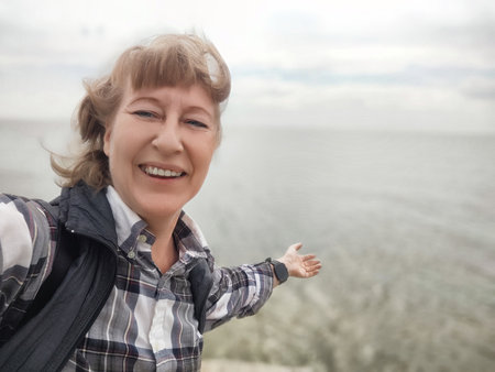 Middle-aged woman with a cheerful smile takes a selfie at the beach, sharing her joyful travel experience during a day of adventure.の写真素材