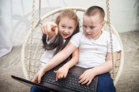 Two children engage in playful activity on a laptop while sitting on a cozy swing indoors in a bright settingの写真素材