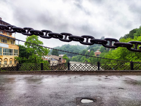 Heavy chain links dominate the foreground while a river winds through a tranquil landscape filled...