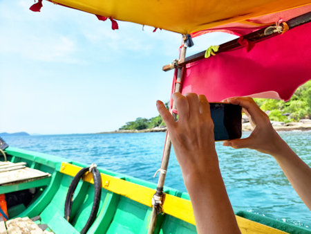 A person is taking a picture with a smartphone while enjoying a boat ride on sparkling blue waters beneath a vibrant canopy.の写真素材