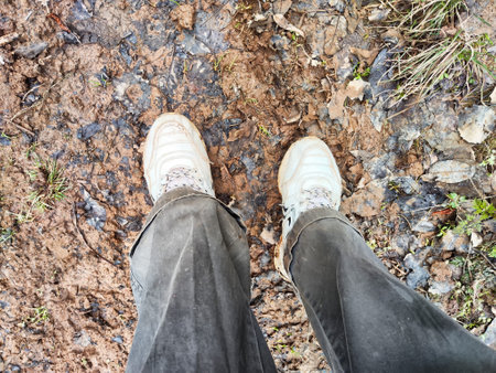 A person stands in muddy ground wearing sneakers, surrounded by earthy tones and plant life on a cloudy day in the outdoors.の写真素材