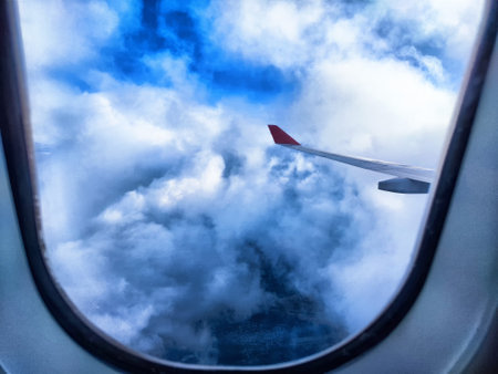 Cloud-filled sky is visible from airplane window, showcasing the wing against vibrant blue and white hues.の写真素材