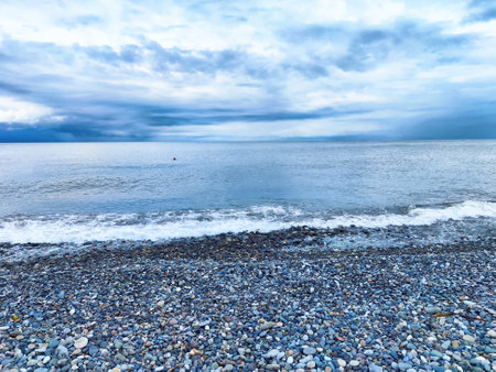 Pebbles line the shore as gentle waves lap against the beach under a cloudy dusk sky, creating a tranquil coastal atmosphere.の写真素材