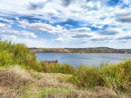 Lush greenery surrounds a serene lake with distant hills under a blue sky scattered with clouds. A quaint house sits by the shore.の写真素材