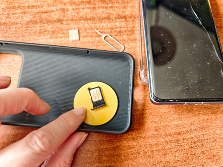 A hand applies a circular sticker to the back of a mobile phone case while a SIM eject tool and a smartphone sit nearby.の写真素材