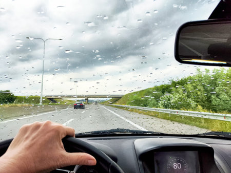 A hand grips the steering wheel of a car driving along a highway during a rain shower, with clouds hanging low and visibility reduced.の写真素材
