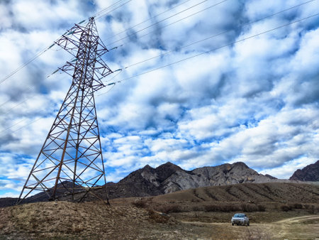 A tall power line tower reaches towards a cloudy sky, overlooking rugged mountains and a quiet dirt road where a vehicle is parked.の写真素材