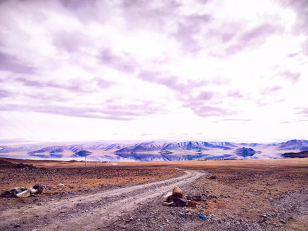 Winding dirt road leads through expansive mountain terrain with a tranquil lake under a cloudy sky, creating a peaceful atmosphere.の写真素材